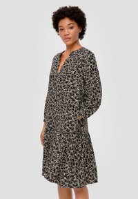 Schwarzes Blumenkleid mit langen Ärmeln, V-Ausschnitt und Seitentaschen. Hat eine fließende Passform und ein kleines weißes Blumenmuster auf ganzer Fläche.