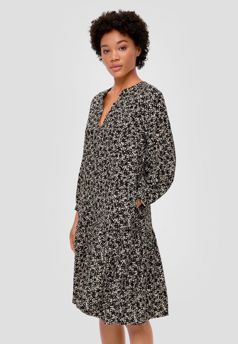 Schwarzes Blumenkleid mit langen Ärmeln, V-Ausschnitt und Seitentaschen. Hat eine fließende Passform und ein kleines weißes Blumenmuster auf ganzer Fläche.