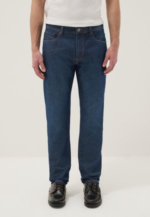 Homme portant un jean bleu foncé à jambes droites, un t-shirt blanc et des chaussures en cuir noires à lacets, debout devant un fond neutre.