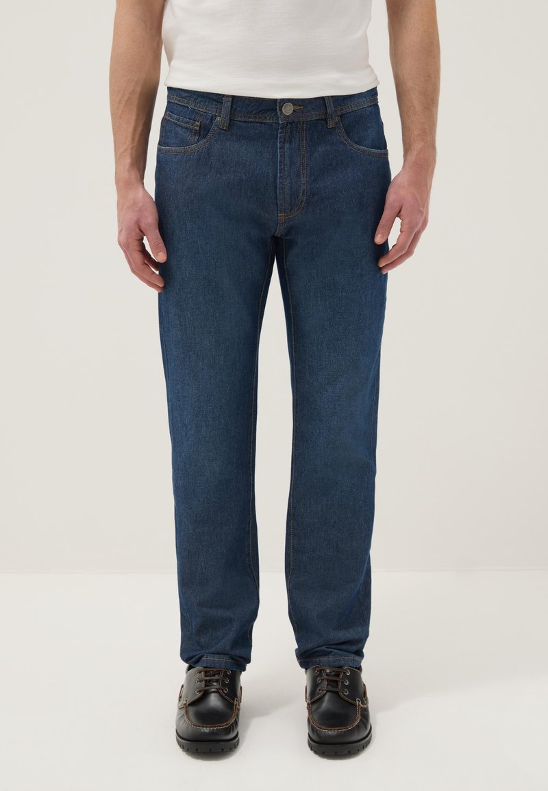 Mann trägt dunkelblaue Jeans mit geradem Bein, weißes T-Shirt und schwarze Leder-Schnürschuhe, steht vor neutralem Hintergrund.