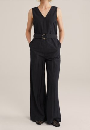 Combinaison noire avec un décolleté en V profond, pantalon à jambe large, ceinture avec une boucle circulaire dorée et poches latérales. Tissu à texture lisse.