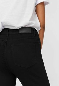 Personne portant un jean noir et un t-shirt blanc, vue de dos de la taille jusqu'au milieu de la cuisse sur un fond uni.