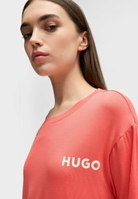 Korallfärgad, lös t-shirt med vid halsringning och korta ärmar, med "HUGO" tryckt i stora vita bokstäver på framsidan.