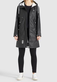 Černý nepromokavý pláštěnka s kapucí lemovanou bílou barvou, s patentními knoflíky, postranními kapsami a logem umístěným poblíž lemu. Nositá přes černé legíny.