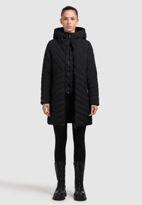 Zwarte doorgestikte lange jas met een capuchon, voorzien van een ritssluiting aan de voorkant, chevronpatroon en zijzakken. Draagt over zwarte leggings en combatlaarzen.