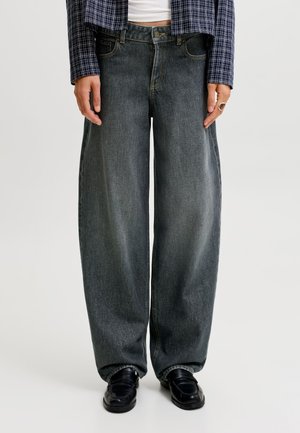 Personne portant un jean gris foncé ample, des mocassins noirs, un haut blanc et une veste à carreaux bleu marine, debout devant un fond uni.