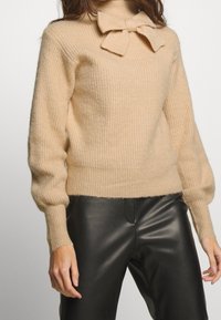 Beige gestrickter Pullover mit gerippter Textur, der ein Schleifendetail am Halsausschnitt und eng anliegende Ärmel aufweist, kombiniert mit schwarzen Lederhosen.