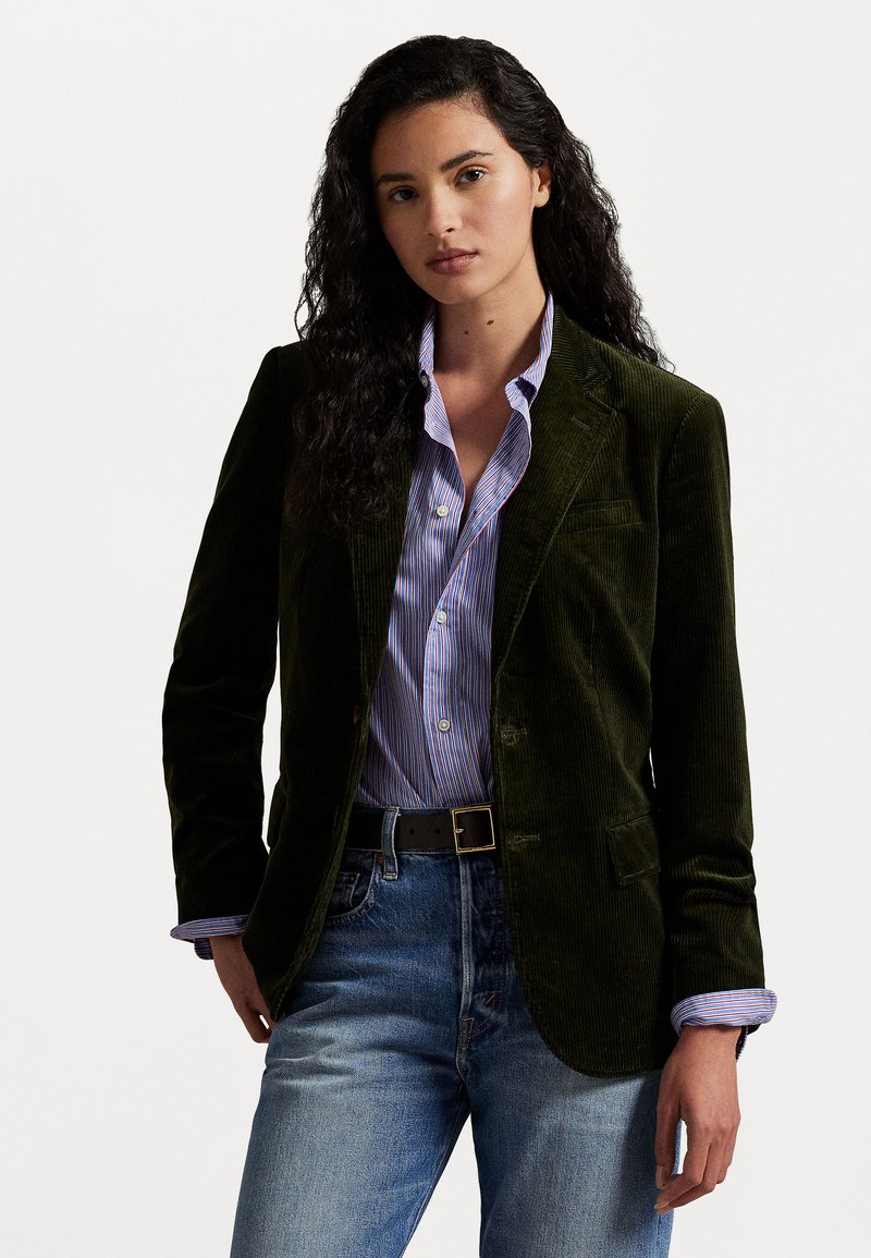 Dark green corduroy blazer with a tailored fit, featuring two front pockets. Worn over a light blue and white striped shirt, paired with blue jeans.
