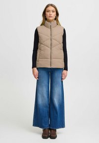 Beige vadderad väst med hög krage och framdragkedja, bärs över en svart långärmad skjorta, ihop med vida blå jeans och bruna stövlar.