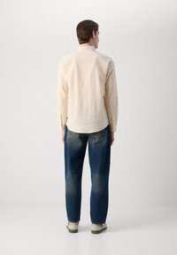 Light beige long-sleeve shirt with a curved hem, paired with loose blue jeans showing a gradient fade and casual footwear in gray.