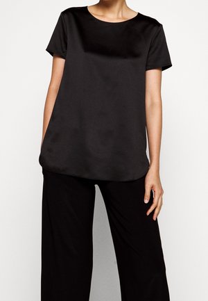 Kobieta ubrana w luźną czarną bluzkę z krótkim rękawem i szerokie czarne spodnie, stojąca na tle jednolitego jasnego tła.