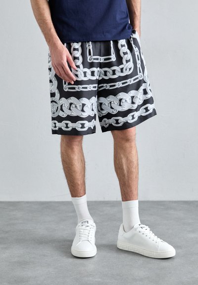 Shorts noirs avec un motif de chaîne blanche, taille élastique et poches latérales, associés à des baskets blanches et des chaussettes mi-mollet.