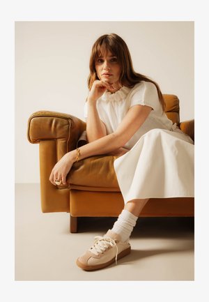 Femme aux cheveux bruns portant un haut en maille blanc, une jupe blanche et des baskets beiges, assise sur un fauteuil en cuir marron, reposant son menton sur sa main.