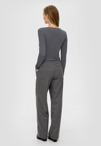 Vrouw die met de handen in de zakken staat, gekleed in een aansluitend grijs longsleeve-top en losse grijze broek, van achteren weergegeven tegen een effen achtergrond.