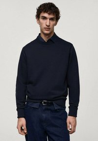 Námornícky modrý pulóver s okrúhlym výstrihom a rebrovanými manžetami, vyrobený z mäkkej tkaniny, je vrstvený cez košeľu s golierom. Má uvoľnený strih.