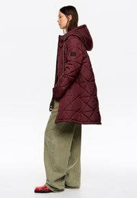 Marszczona bordowa puchowa kurtka z kapturem, z naszywką z logo na rękawie. W zestawie z oliwkowozielonymi szerokimi spodniami i czerwonymi butami w wzory.