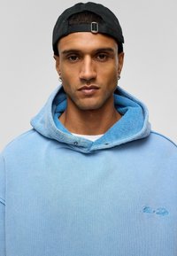 Lichtblauwe denim hoodie met een contrasterende blauwe fleecelaag in de capuchon, drukknopen details en geborduurd logo op de borst. Zwarte pet gedragen.