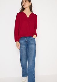 Rød bluse med lange ærmer, V-hals og knaplukning, parret med brede blå jeans i denim. Modellen har hvide sneakers på.