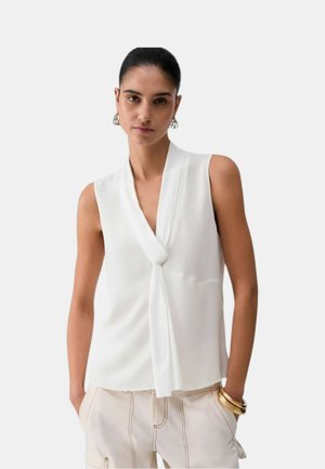 Donna con capelli scuri raccolti indossa una camicetta bianca senza maniche con nodo frontale e pantaloni chiari con dettagli di cucitura.