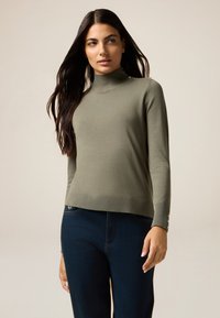 Maglione aderente a collo alto verde oliva realizzato in morbido materiale; presenta un orlo a costine e dettagli con bottoni ai polsini. Abbinato a jeans scuri.