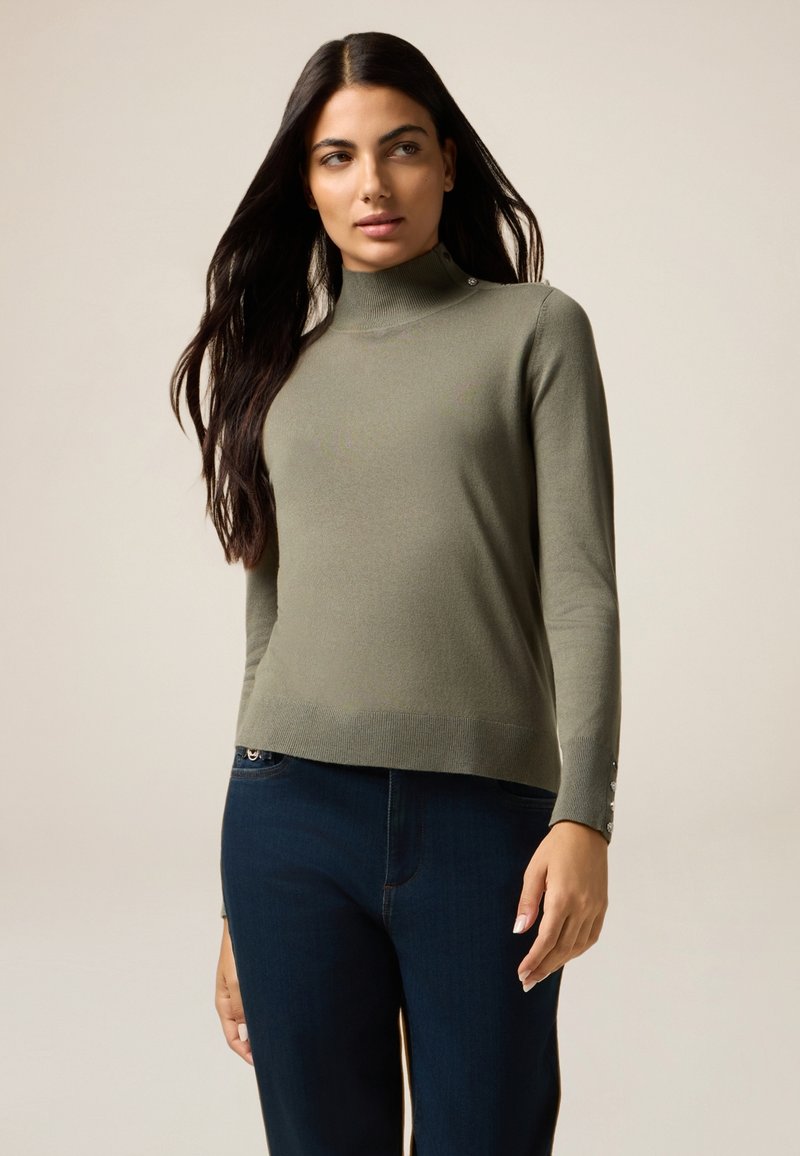 Maglione aderente a collo alto verde oliva realizzato in morbido materiale; presenta un orlo a costine e dettagli con bottoni ai polsini. Abbinato a jeans scuri.