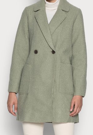 Femme portant un manteau croisé texturé vert clair par-dessus un pull à col roulé blanc et un pantalon crème.