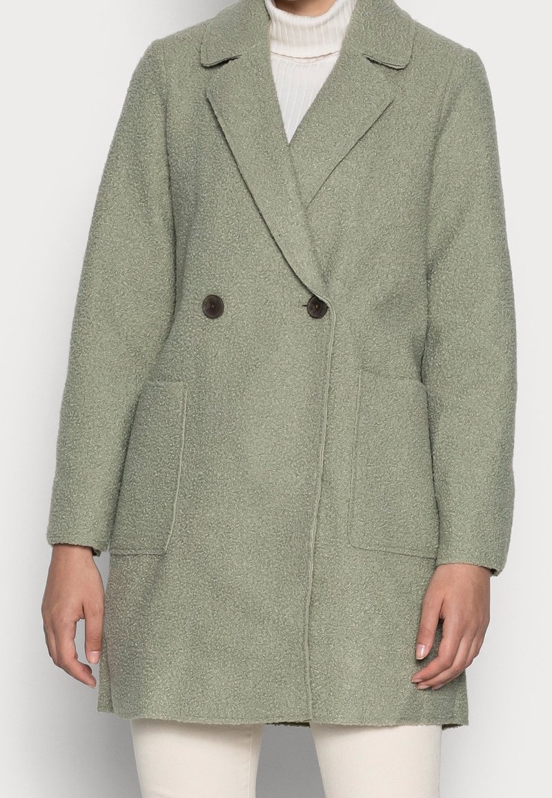 Femme portant un manteau croisé texturé vert clair par-dessus un pull à col roulé blanc et un pantalon crème.
