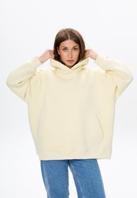 Felpa oversized color crema con tasca a marsupio, cappuccio con lacci e polsini a coste, abbinata a jeans in denim blu.