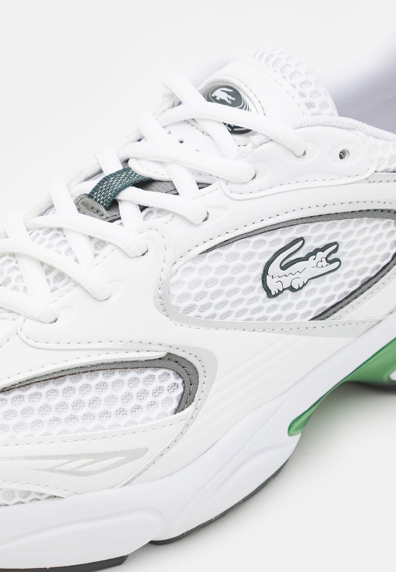 Sneaker atletica bianca con pannelli in rete, accenti grigi e una suola verde. Presenta una chiusura con lacci e un piccolo logo lato.