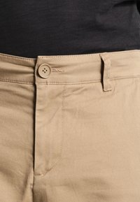 Beżowe spodnie z zapięciem na guziki, z gładkiej, teksturowanej tkaniny z przeszyciami. Pas wyposażony w szlufkę na pasek.