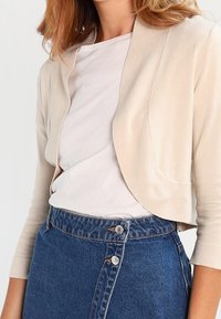 Žena v béžovém zkráceném kardiganu přes bílé tričko a v modré riflové sukni s vysokým pasem, která má vpředu dva knoflíky.