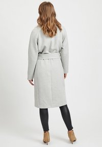 Lang grå frakke med bælte, med brede ærmer og lige snit, parret med sorte leggings og beige ankelstøvler.