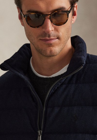 Vyras dėvi vėžlio kiauto spalvos akinius nuo saulės ir tamsiai mėlyną striukę su užtrauktuku, užsivilkęs tamsų megztinį su matoma balta apykakle.