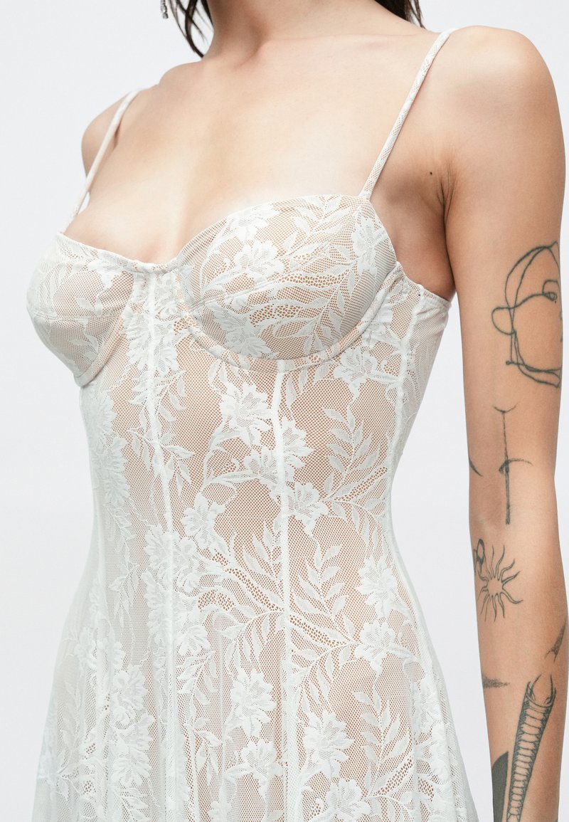 Femme portant une robe en dentelle blanche transparente à motif floral et bretelles fines, montrant des tatouages sur son bras droit sur un fond uni.
