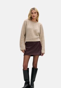 Beżowy sweter na drutach z dużymi rękawami, zestawiony z bordową mini spódnicą ze skóry i czarnymi kozakami za kolano, na neutralnym tle.