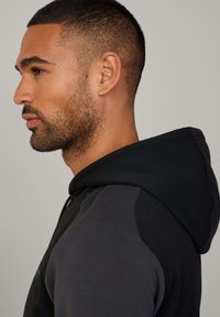 Sudadera negra con cuello alto, textura suave y detalles de cremallera lateral. Presenta un diseño en dos tonos con acentos en gris oscuro en contraste.