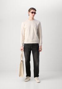 Felpa beige chiaro, jeans scuri, sneakers bianche. Il modello tiene una borsa a tracolla beige chiaro. Occhiali da sole circolari completano il look.