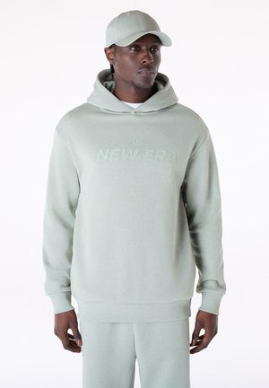 Mann trägt einen hellgrünen Hoodie mit der Aufschrift „"NEW ERA"“, passende Jogginghose und eine Kappe, steht vor einem einfachen, hellen Hintergrund.