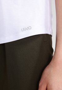 Bílý bavlněný tričko s volným střihem, zdobené názvem značky z kamínků na lemu, kombinované s tmavě olivovými kalhotami.