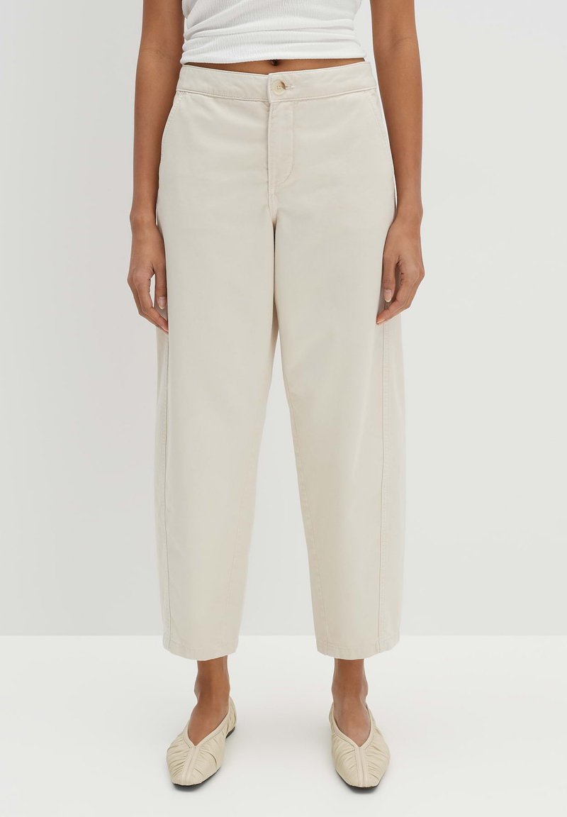 Pantalons beige à jambes larges en coton, avec une fermeture à bouton, des poches avant et une texture lisse. Associés à des chaussures plates de couleur claire.