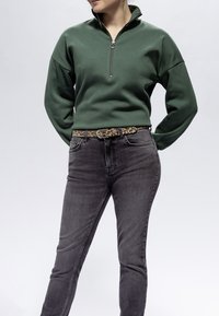 Donna con felpa verde scuro con mezza cerniera, jeans neri e cintura con stampa leopardata, in piedi con le mani dietro la schiena su uno sfondo semplice.