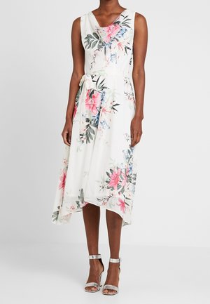 Robe midi blanche sans manches à imprimé floral rose, bleu et vert, avec taille nouée, et sandales à talons hauts argentées avec bride à la cheville.