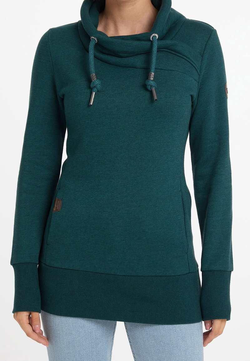 Femme portant un sweat à capuche à manches longues vert foncé avec cordons, poignets et ourlet côtelés, associé à un jean bleu clair, debout sur fond blanc.