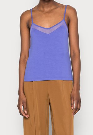 Woman wearing a sleeveless purple camisole with sheer panel at the neckline and loose brown pants, standing against a plain background.