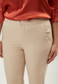 Pantaloni beige aderenti con texture liscia, tasche frontali e chiusura con zip, abbinati a un top in satin rosso.