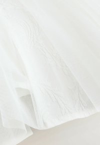 Tessuto bianco trasparente con delicato ricamo floreale sovrapposto a un tessuto bianco solido, morbido e leggermente traslucido.