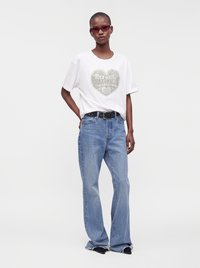 Person wearing red sunglasses, white t-shirt with heart graphic, blue high-waist jeans, black belt, and pointed black shoes, standing on white background.
