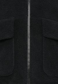 Svart fleecejacka med textur, central silverfärgad dragkedja och två fickor med lock.