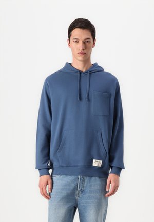Blue cotton hoodie with a front pocket, drawstring hood, and ribbed cuffs. Features a small label at the hip. Paired with light blue jeans.