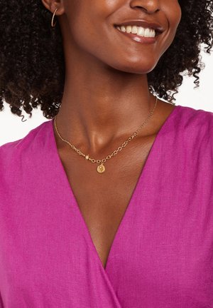 Collier en or avec un pendentif circulaire et une accent étoile, porté avec un haut enroulé rose vif en tissu texturé.